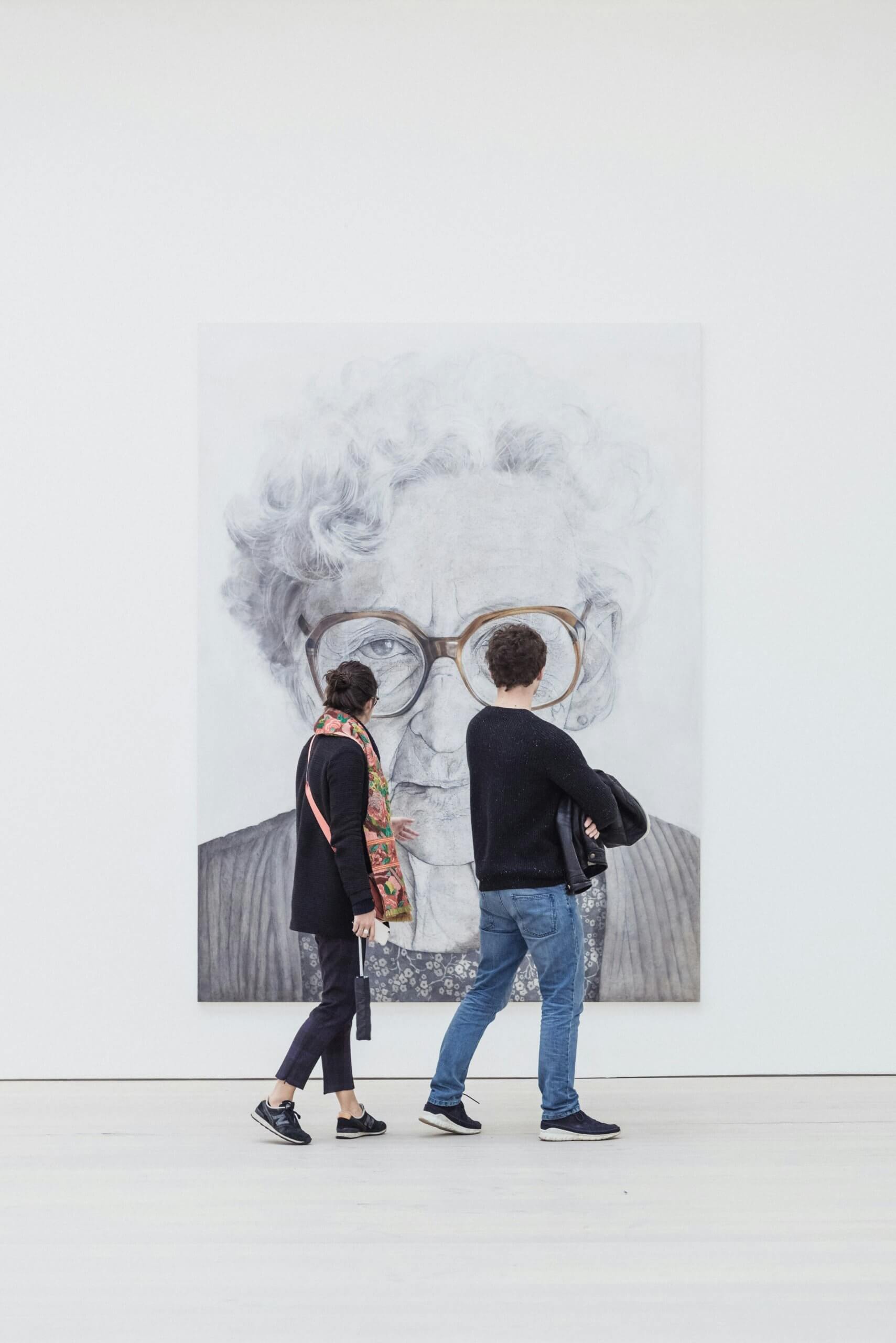 Photo of two people at a light-filled museum viewing a large portrait of a woman with glasses.
