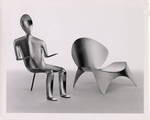 Black and white photograph of two metallic chairs, each constructed from a single sheet of aluminum, one of which is shaped like a seated human form.