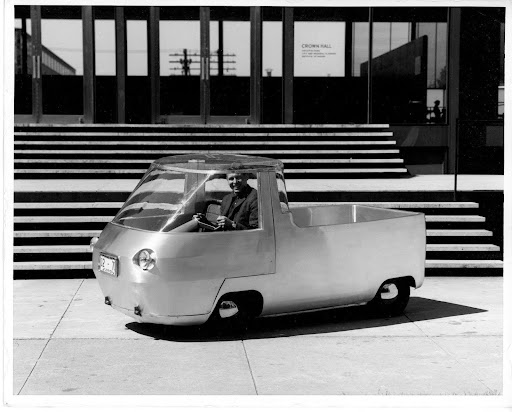 Black and white photograph of a white man sitting inside an old-fashioned car parked in front of a staircase leading up to the Crown Hall building at IIT.