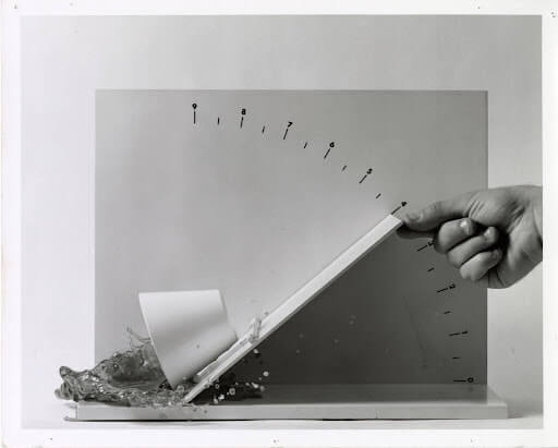 Black and white photograph of a teacup experiment. A plain, white teacup is balanced on a flat surface. A hand lifts the right edge of the surface to a 40 degree angle, at which point the teacup tips toward the left and spills the liquid inside. The photograph captures the moment at which the teacup topples and the liquid spills out of the cup.